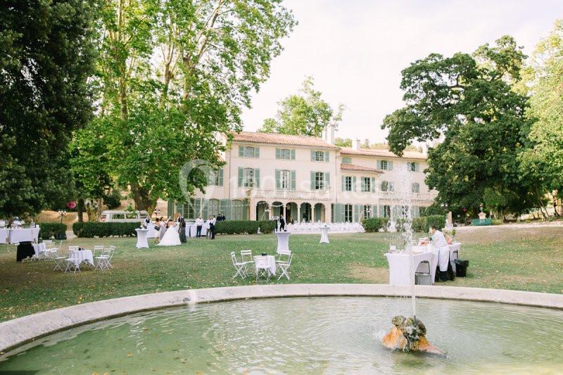 Jardin avec fontaine au premier plan, tables dressées et invités devant une grande maison de style provençal.