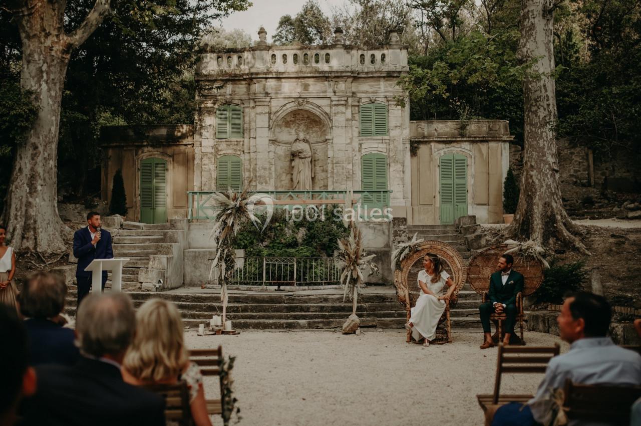 Cérémonie en plein air devant une façade ancienne, avec des invités assis et un orateur debout à un pupitre.
