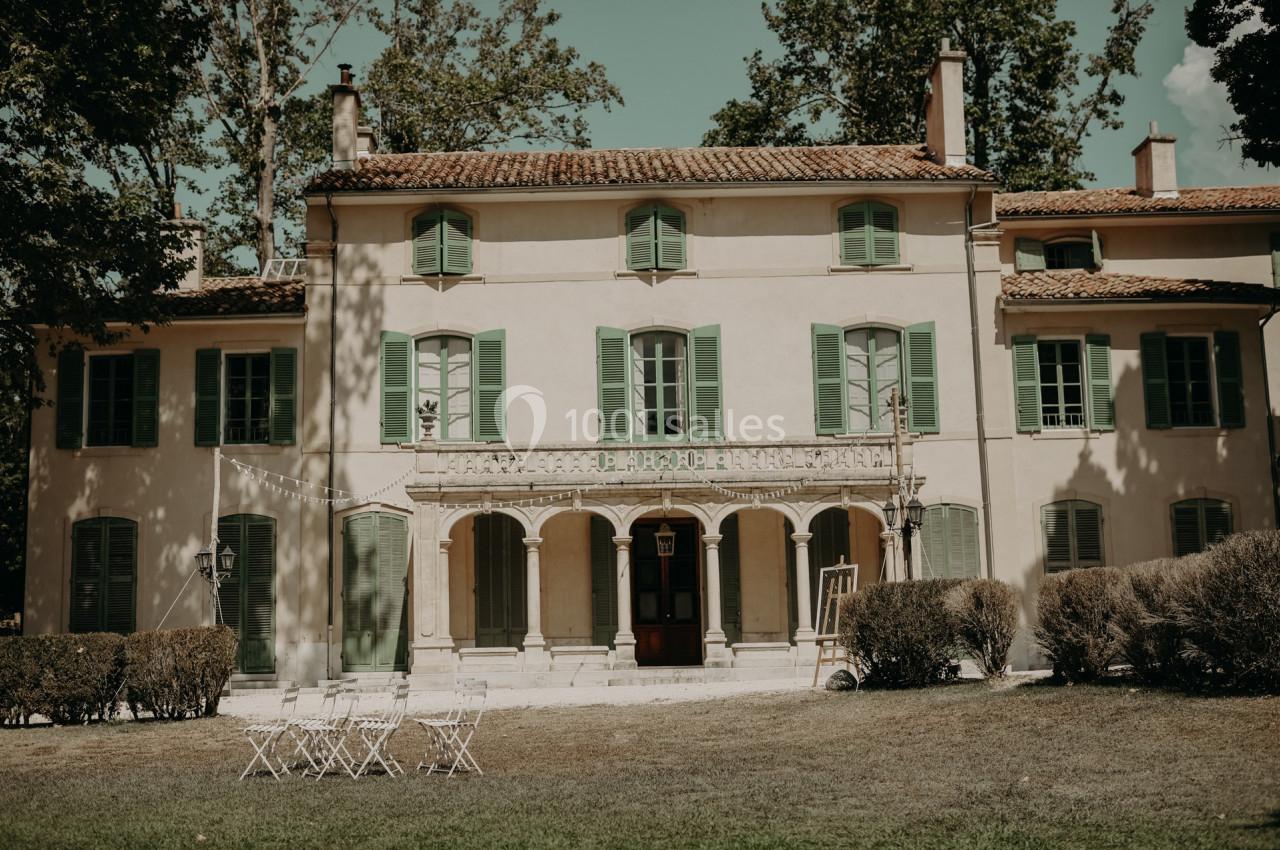 Façade d'une grande maison ancienne avec volets verts, entourée d'arbres et d'un jardin avec des chaises blanches.