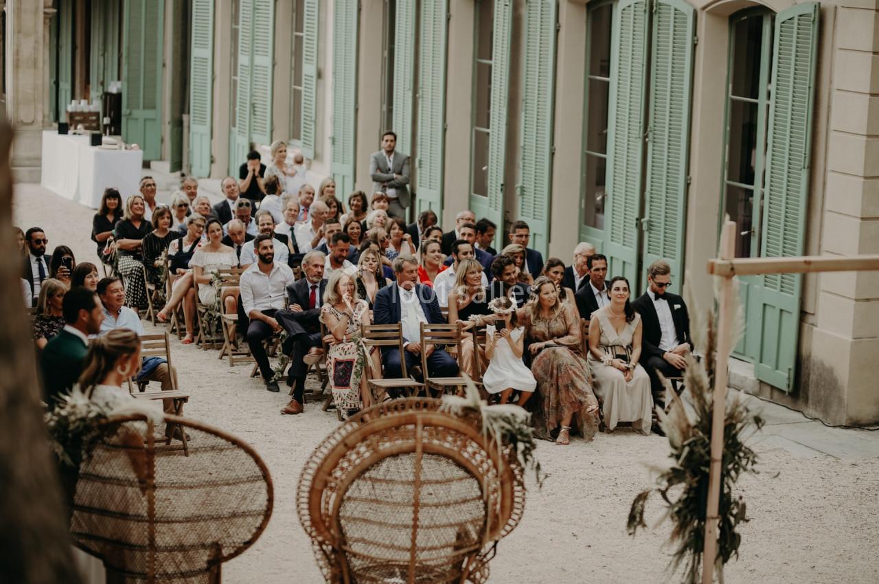 Invités assis en extérieur lors d'une cérémonie, entourés de bâtiments aux volets verts et de décorations naturelles.