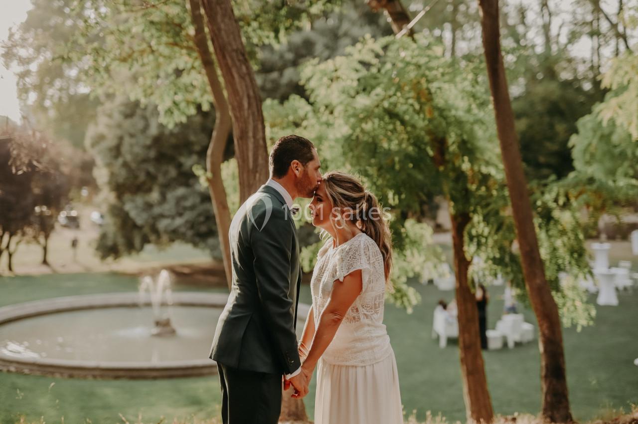Un couple en tenue de mariage se tient la main dans un parc verdoyant près d'une fontaine.