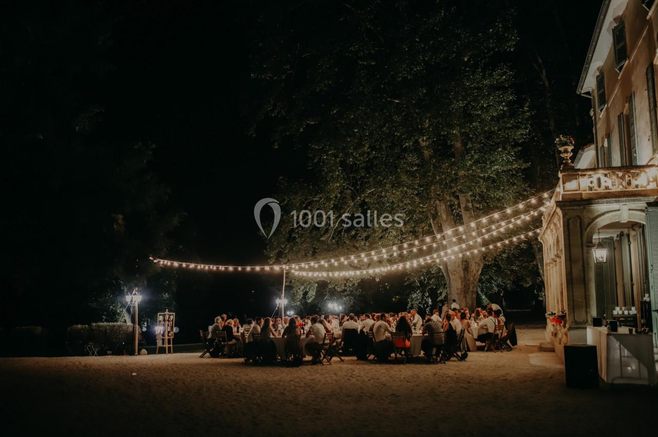 Groupe de personnes dînant en plein air sous des guirlandes lumineuses, près d'un bâtiment éclairé, de nuit.