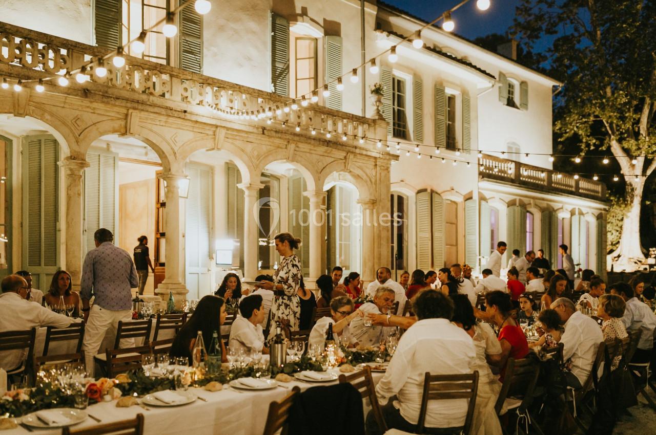 Dîner en plein air avec de longues tables dressées devant une grande maison illuminée, sous des guirlandes lumineuses.