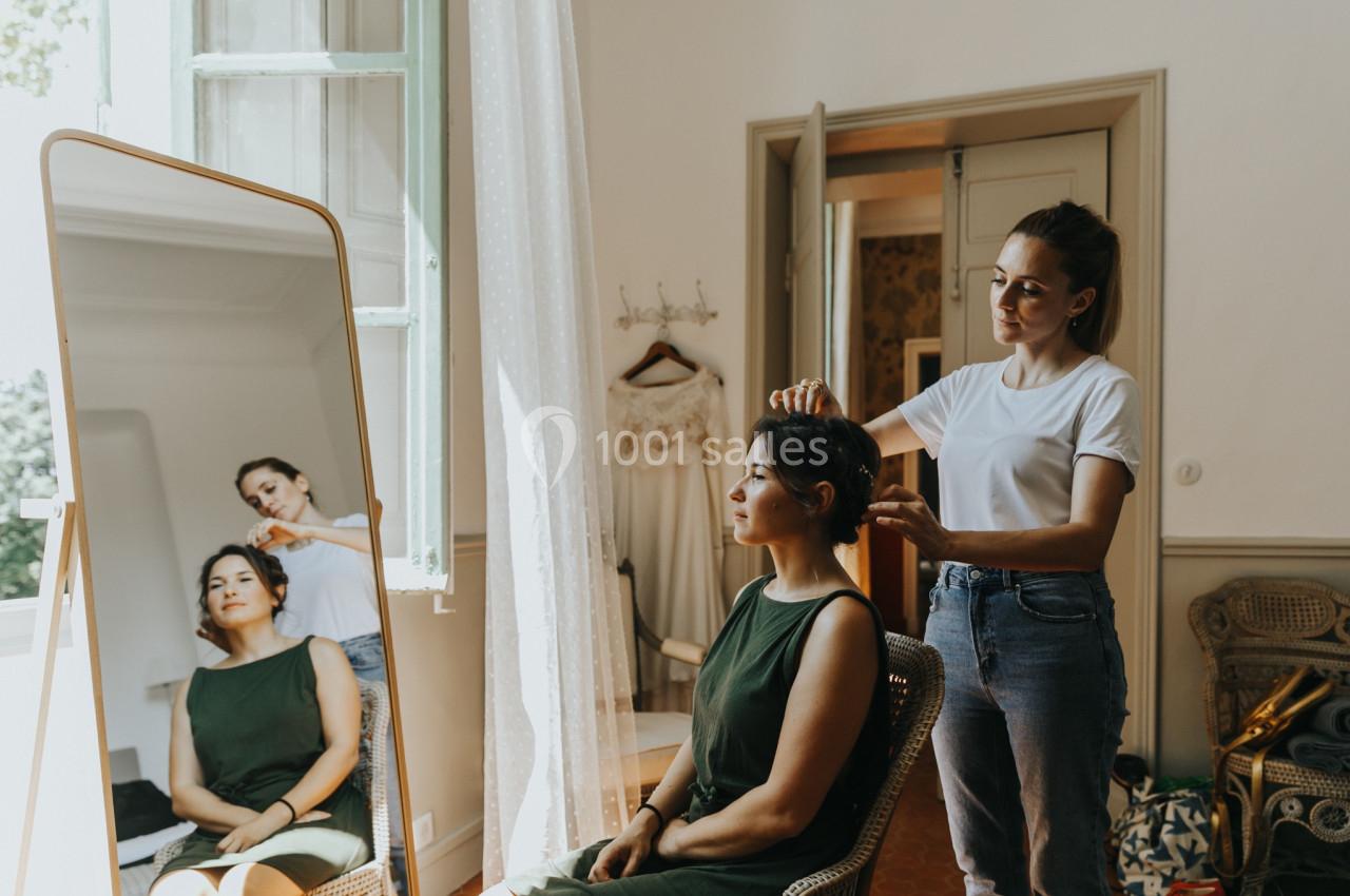 Une femme assise se fait coiffer par une autre femme dans une pièce lumineuse avec un miroir et une robe accrochée au mur.