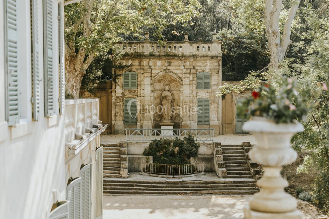 Statue en pierre dans un jardin ancien entouré d'arbres, avec des escaliers et une architecture classique en arrière-plan.