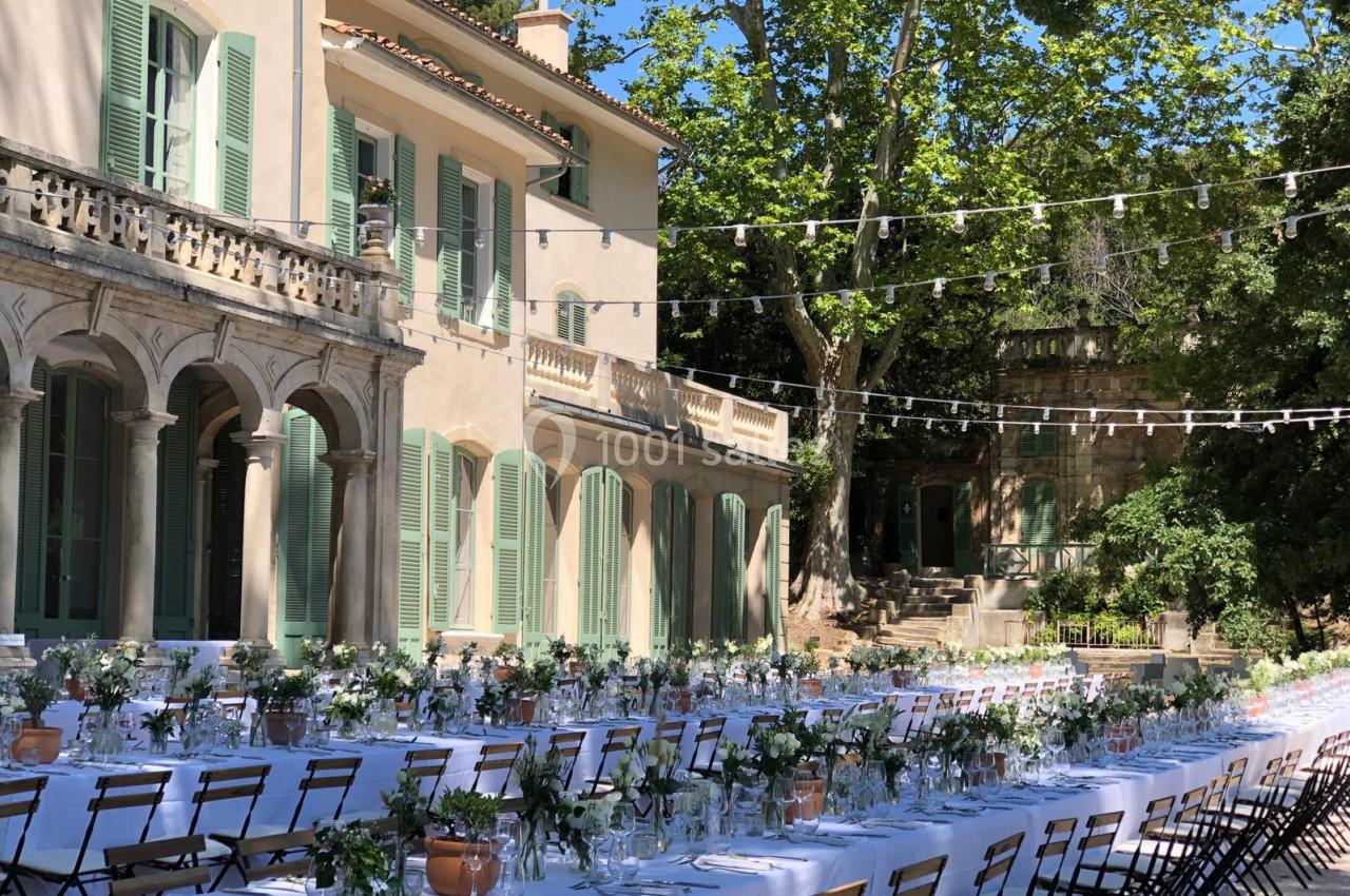 Tables dressées pour un événement en plein air devant une grande maison avec volets verts et guirlandes lumineuses…