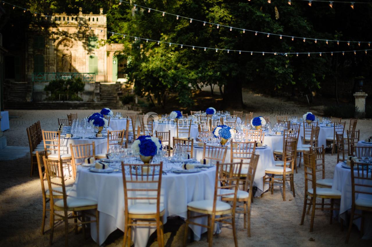 Tables rondes décorées de nappes blanches et de bouquets bleus, disposées en extérieur sous des guirlandes lumineuses.