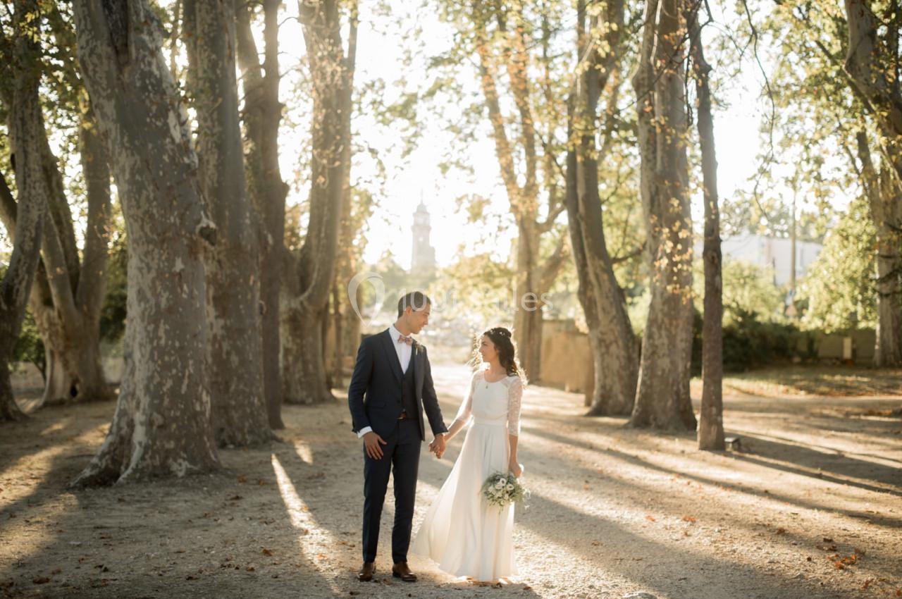 Un couple en tenue de mariage se tient la main dans une allée bordée d'arbres, baignée par une lumière douce.