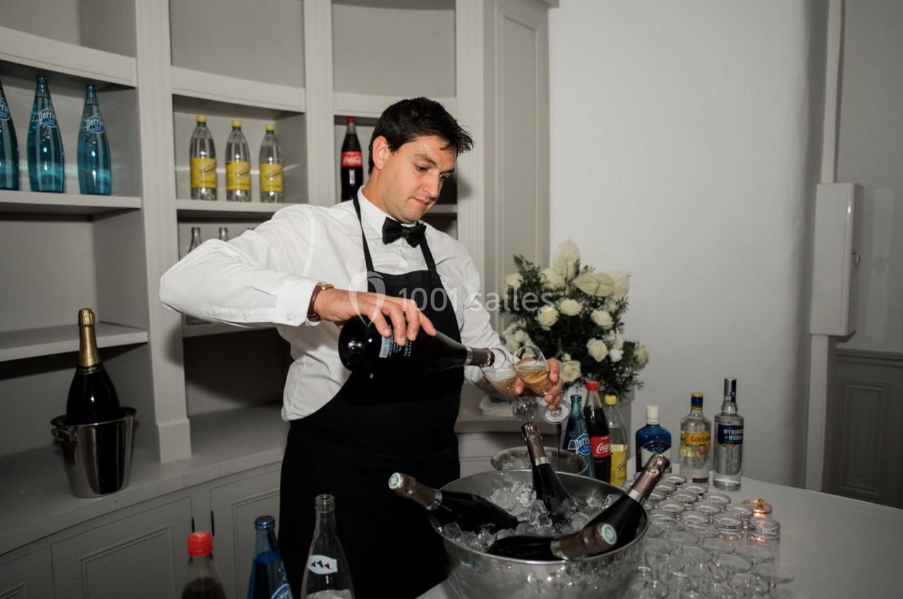 Un serveur en chemise blanche et tablier noir verse du champagne dans un verre devant un bar avec des bouteilles.