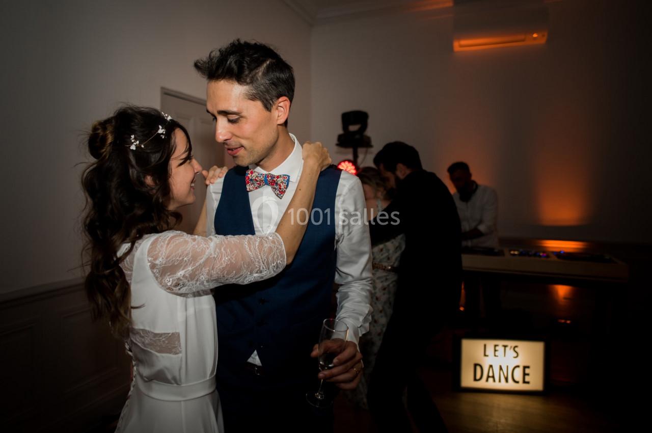 Un couple danse tendrement dans une salle faiblement éclairée, avec une pancarte lumineuse ’Let's Dance’ en arrière-plan.