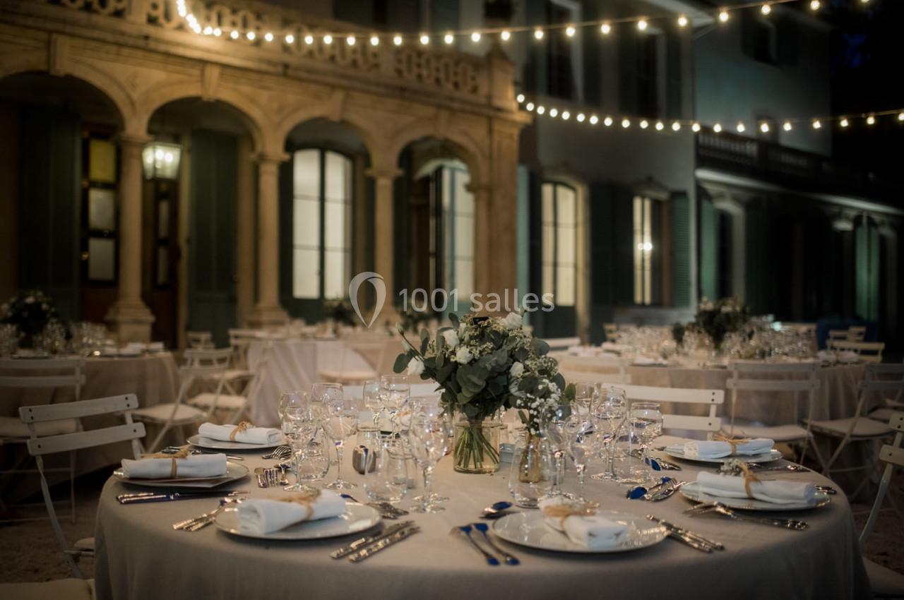 Tables élégamment dressées avec vaisselle et fleurs, disposées en extérieur sous des guirlandes lumineuses.