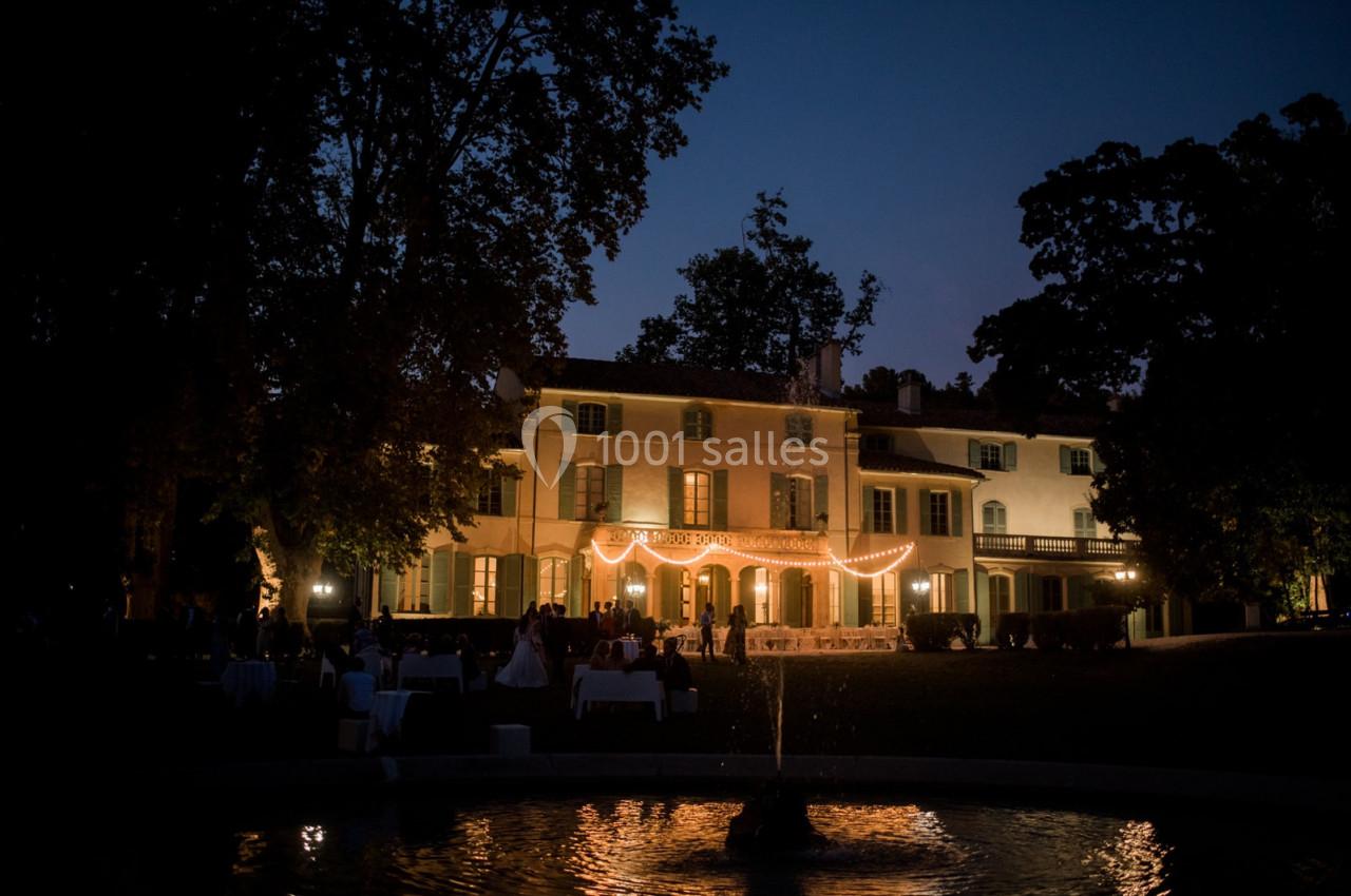 Grande maison éclairée par des guirlandes lumineuses, entourée d'arbres, avec une fontaine au premier plan, de nuit.