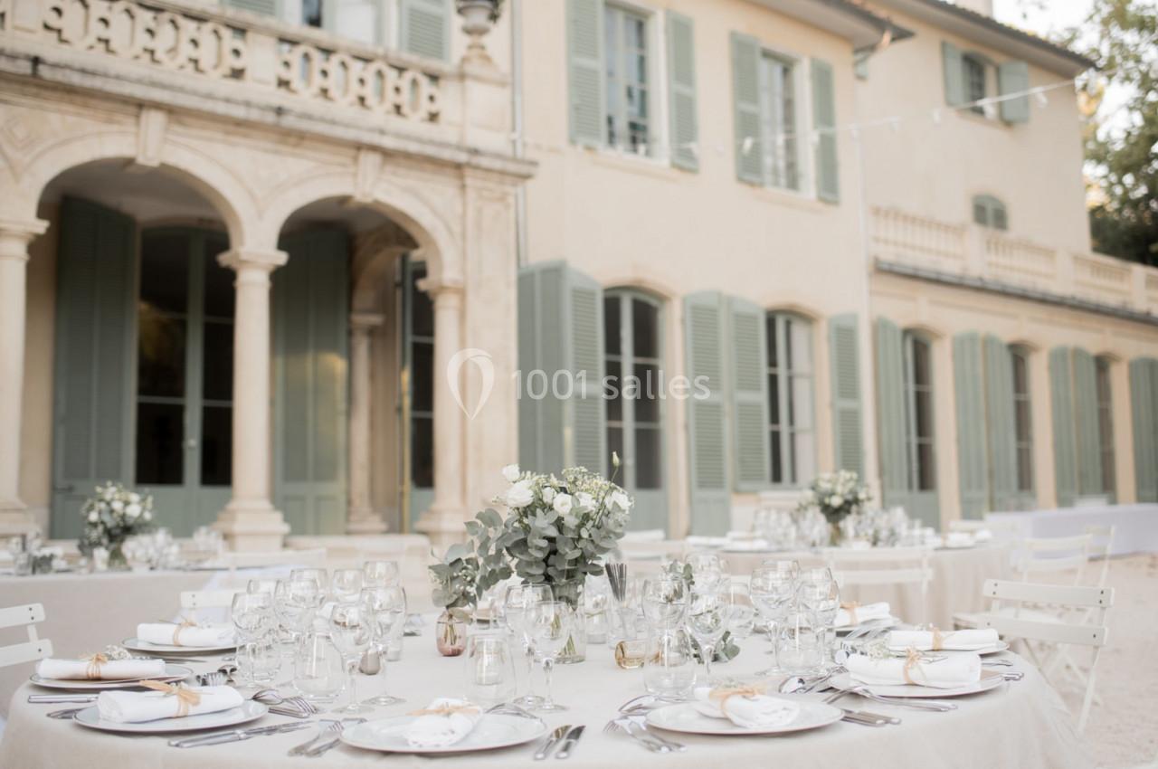 Tables élégamment dressées avec des décorations florales devant une grande bâtisse aux volets verts et arcades en pierre.