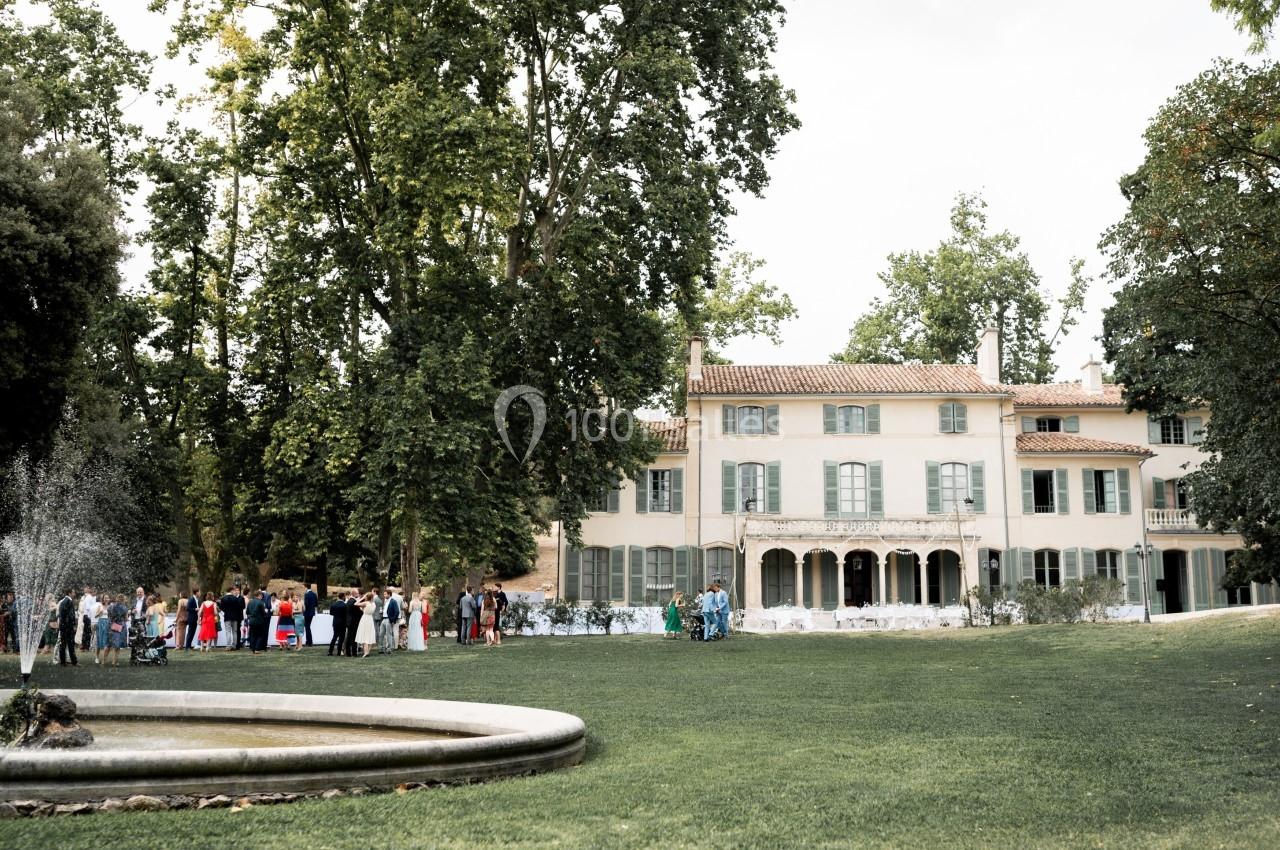 Manoir entouré d'un grand jardin verdoyant avec une fontaine au premier plan et des invités rassemblés à l'extérieur.