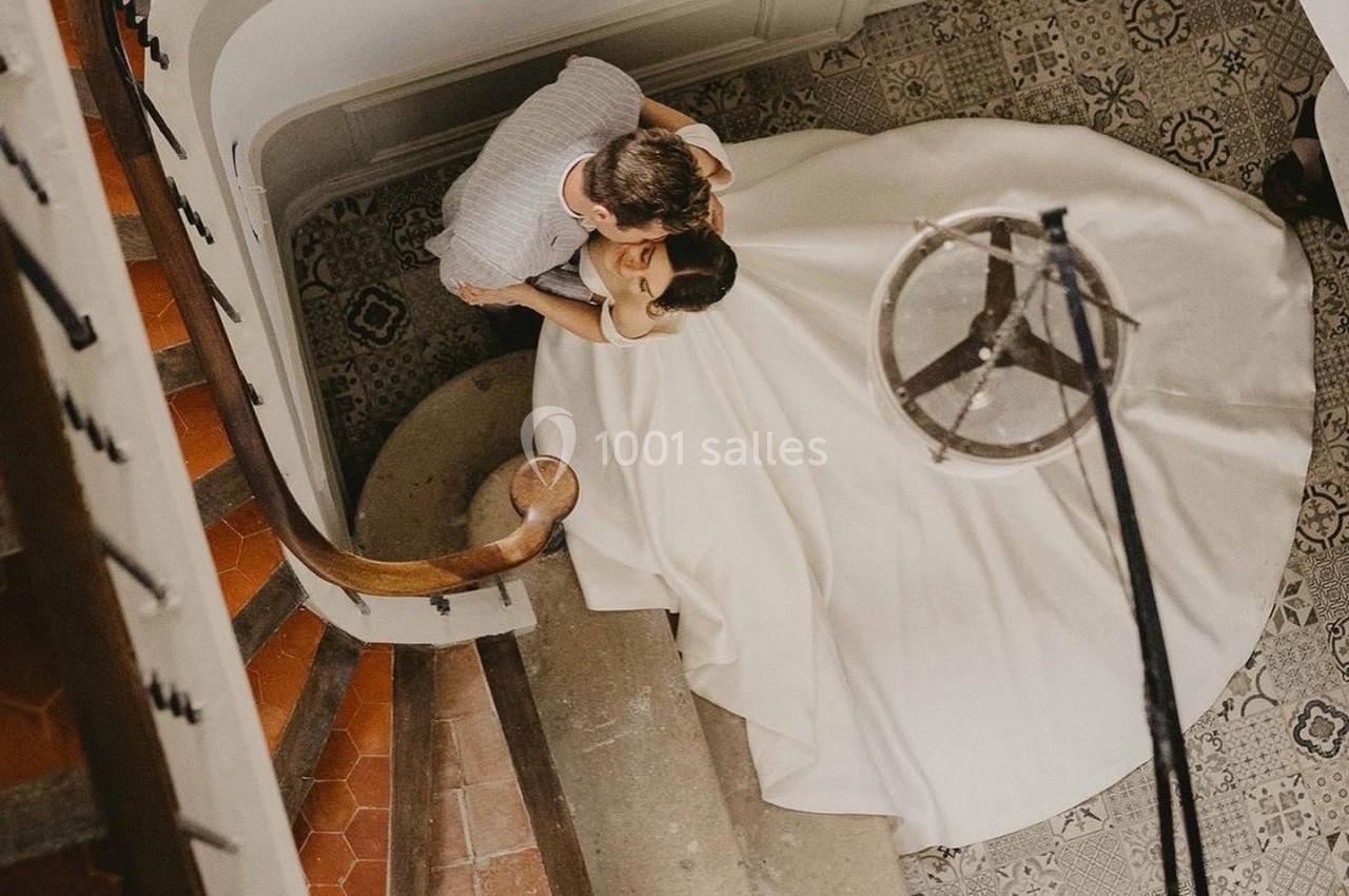 Vue plongeante d'un couple dans un escalier ancien, la femme porte une robe blanche ample.