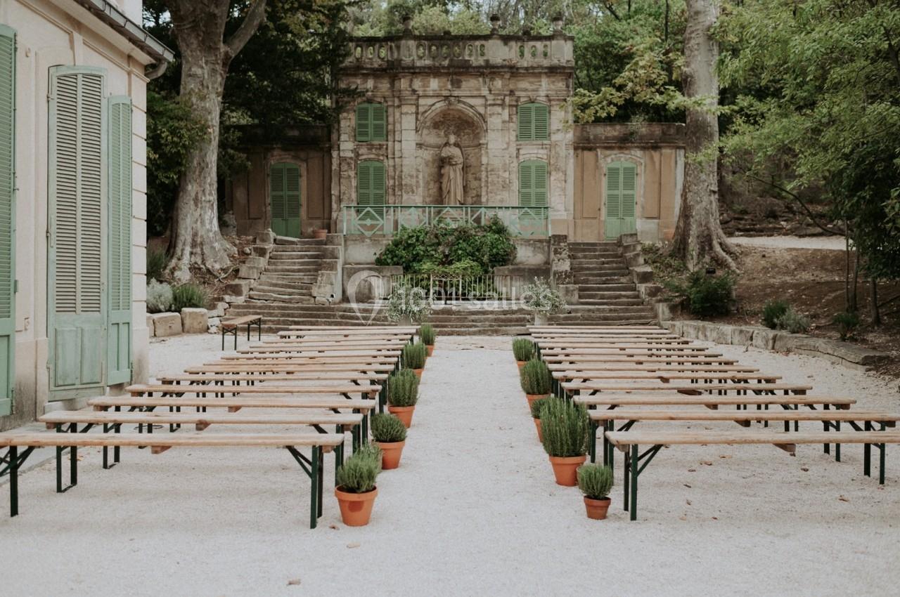 Bancs en bois alignés avec des pots de plantes, face à une façade ancienne dans un jardin ombragé.