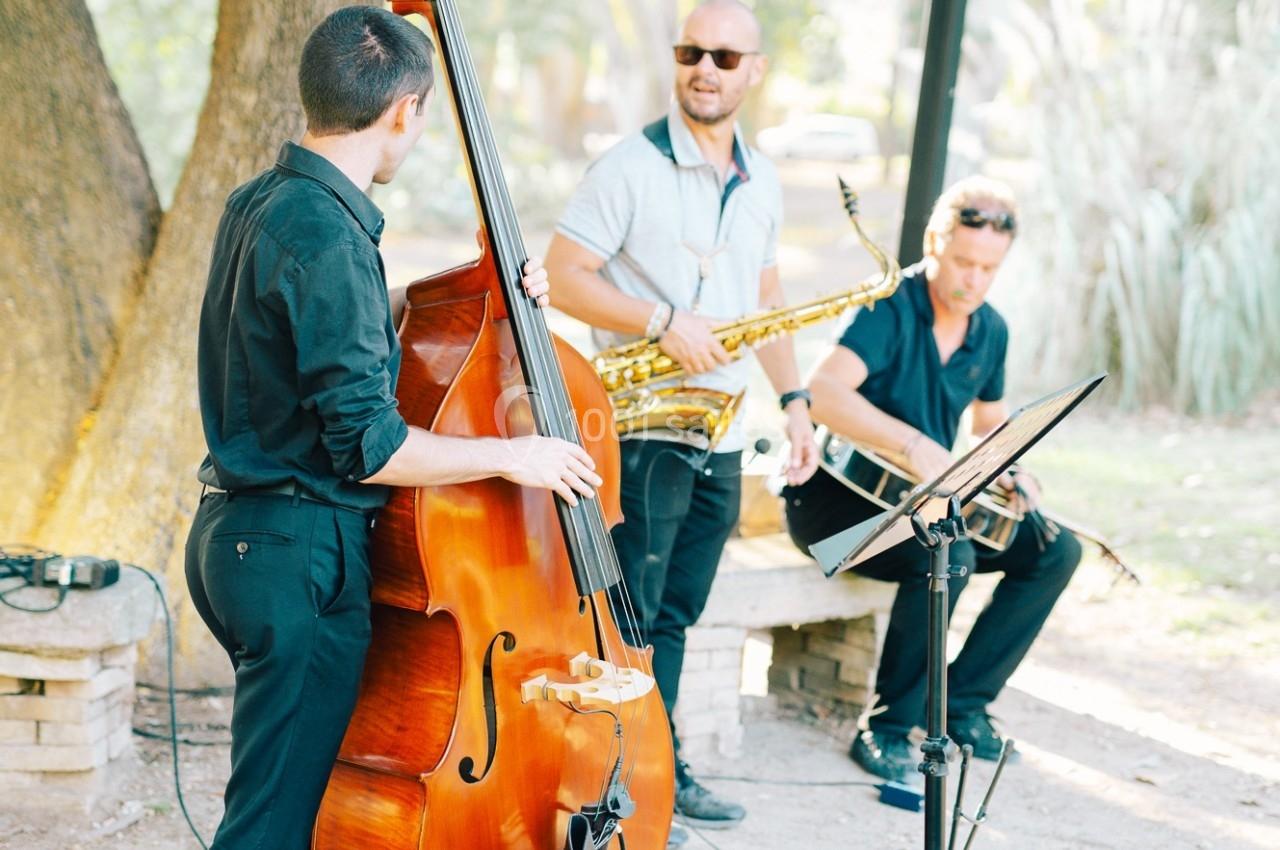 Trois musiciens jouent de la contrebasse, du saxophone et de la batterie en plein air, sous des arbres.