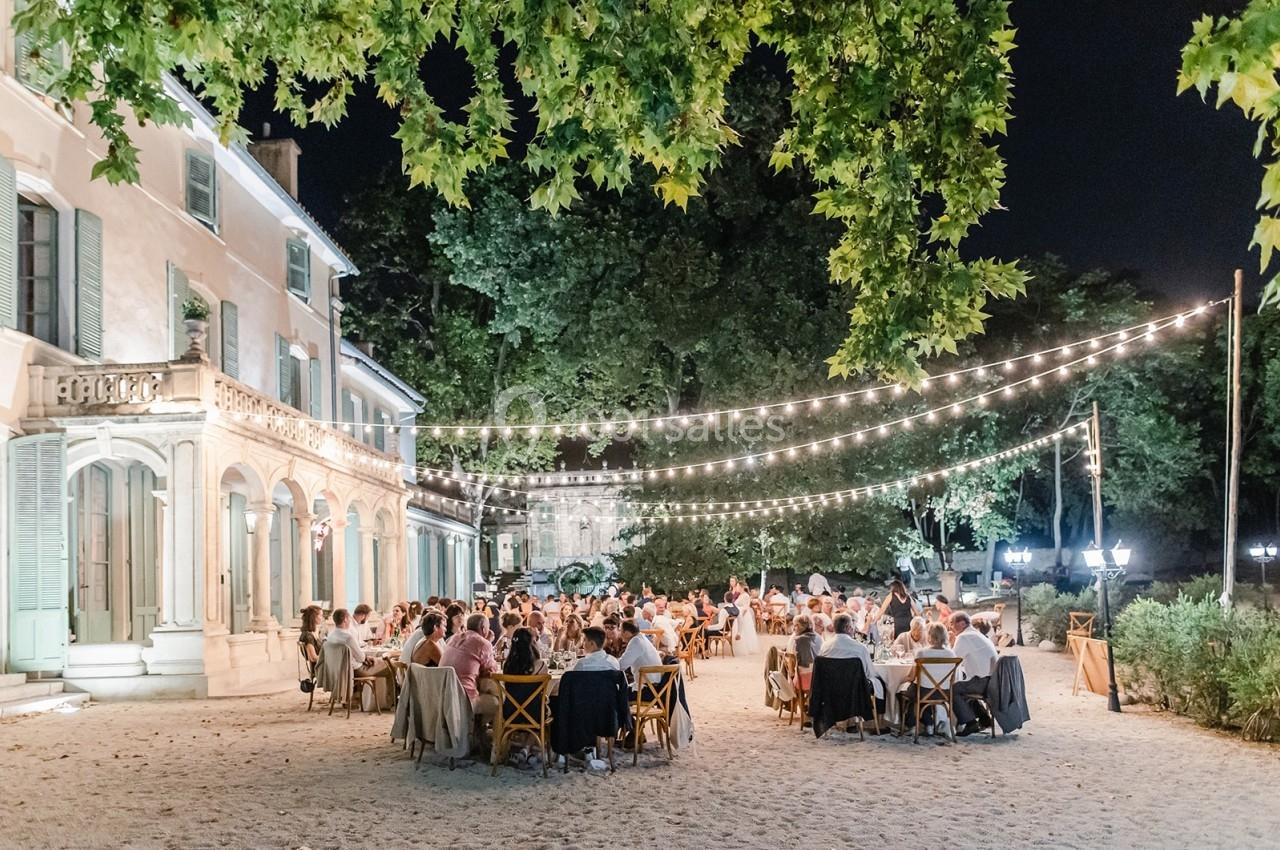 Dîner en plein air sous des guirlandes lumineuses, devant une grande maison entourée d'arbres, de nuit.