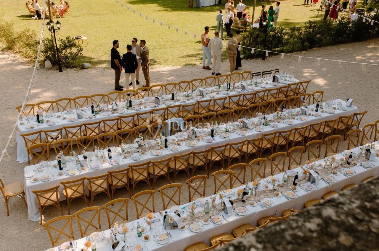 Tables de banquet dressées en extérieur avec nappes blanches, vaisselle et chaises en bois, invités discutant au fond.