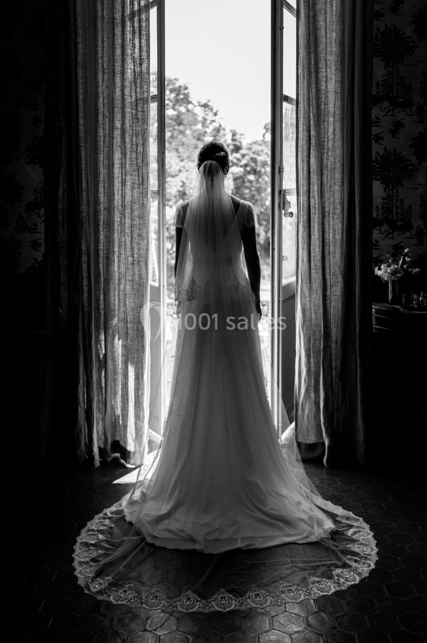 Location salle Marseille 14 (Bouches-du-Rhône) - Bastide de Toursainte #89 Une mariée en robe blanche et voile vue de dos, debout devant une porte-fenêtre ouverte sur un extérieur lumineux.
