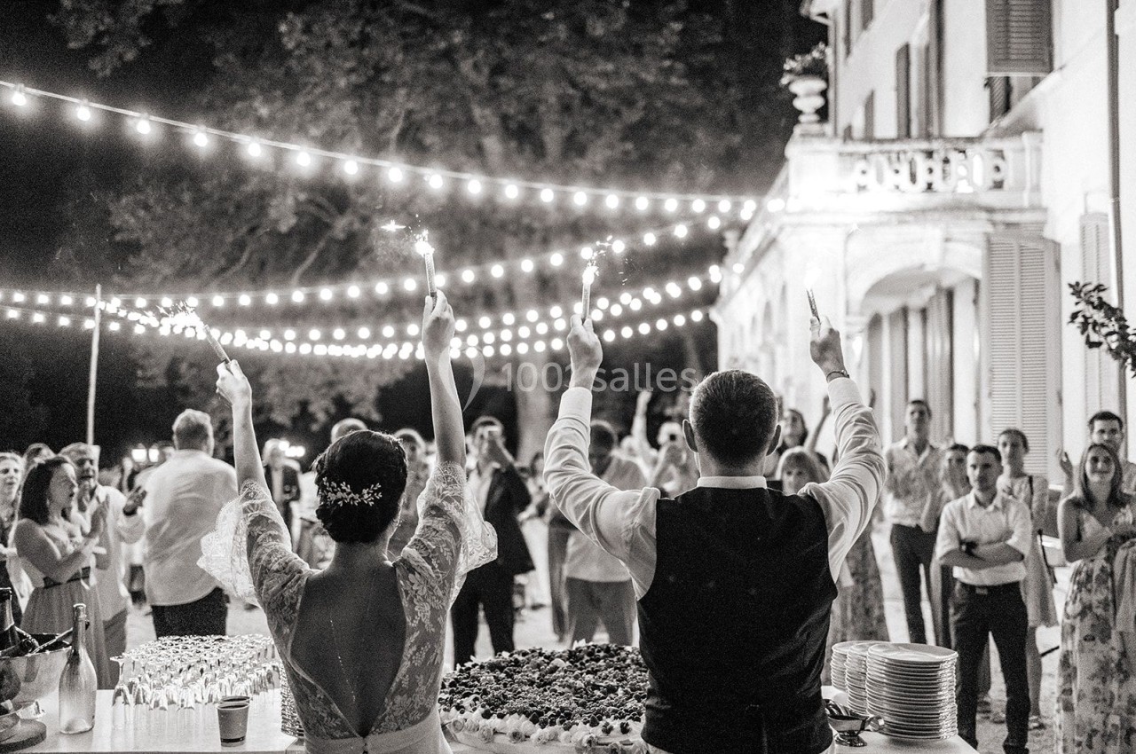 Location salle Marseille 14 (Bouches-du-Rhône) - Bastide de Toursainte #96 Un couple de mariés de dos lève des cierges magiques devant des invités lors d'une fête en extérieur éclairée.