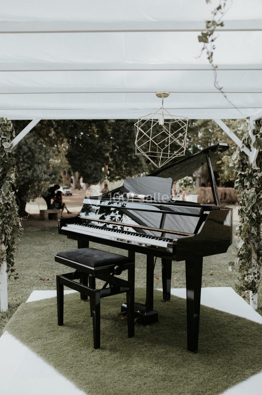 Location salle Marseille 14 (Bouches-du-Rhône) - Bastide de Toursainte #98 Piano à queue noir avec tabouret, installé sur une pelouse sous une pergola décorée de feuillage.