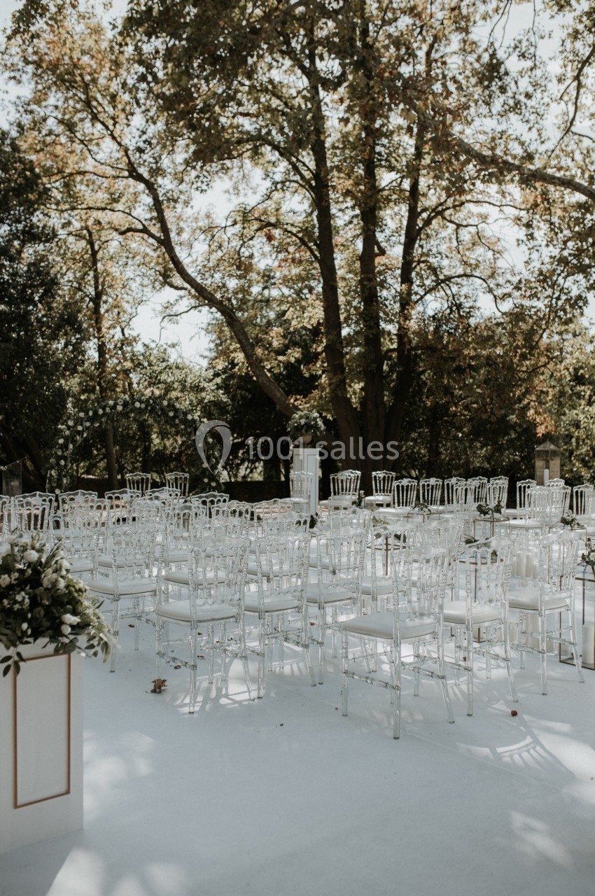 Location salle Marseille 14 (Bouches-du-Rhône) - Bastide de Toursainte #99 Chaises transparentes disposées en rangées sur une allée blanche, entourées d'arbres dans un cadre extérieur.
