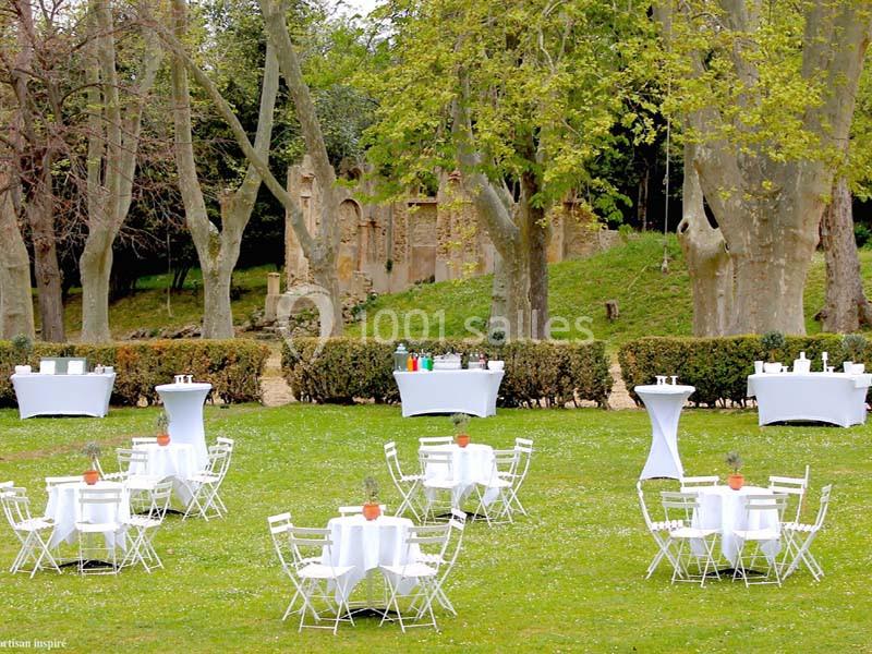 Tables blanches dressées avec chaises sur une pelouse, entourées d'arbres et de haies, dans un cadre extérieur.