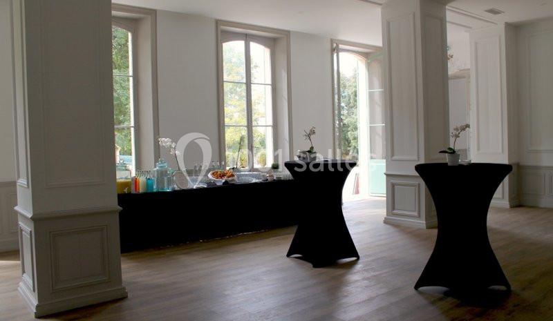 Salle lumineuse avec grandes fenêtres, tables hautes noires et buffet comprenant boissons et collations.