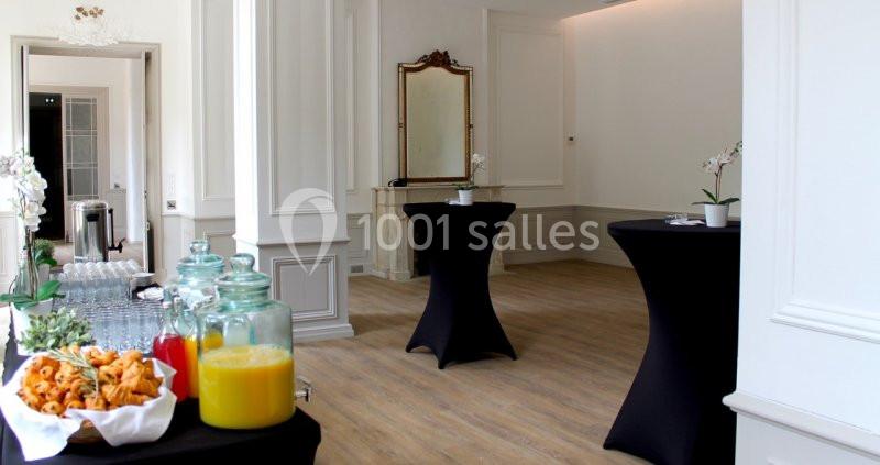 Salle lumineuse avec tables hautes noires, buffet de boissons et snacks, décorée de miroirs et d'orchidées blanches.