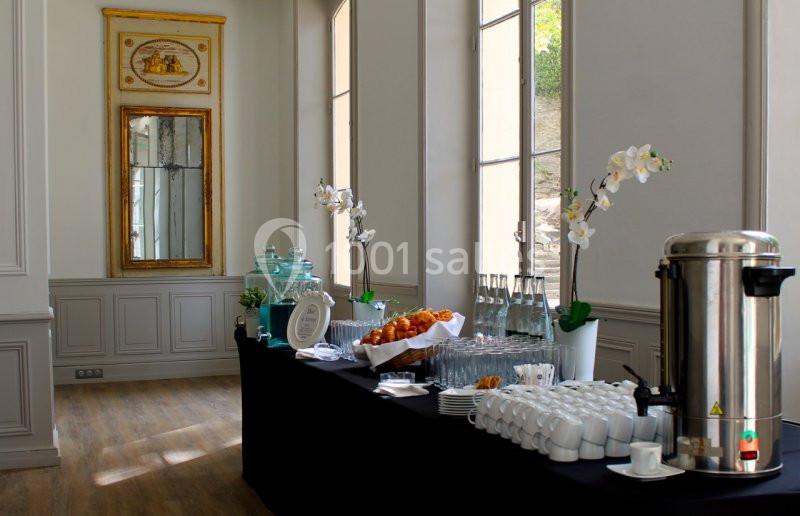 Table de buffet avec boissons, viennoiseries, tasses et décorations florales dans une salle lumineuse au style classique.