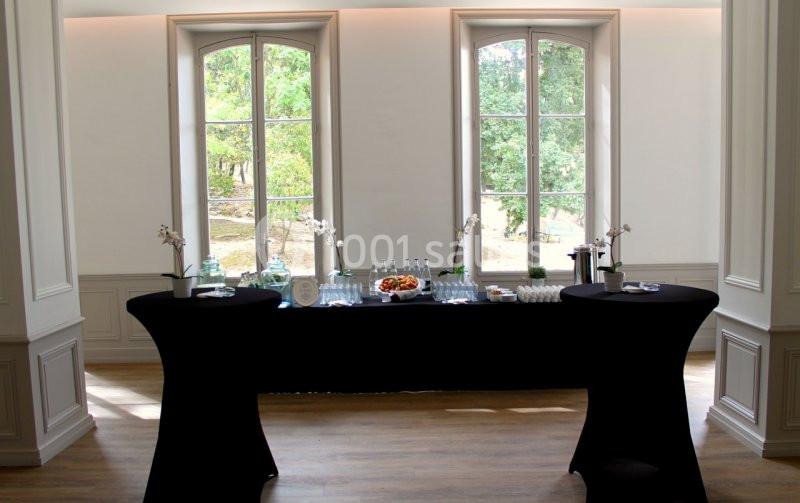 Table de réception avec nappes noires, boissons, verres et plateau de fruits, devant des fenêtres donnant sur un jardin.