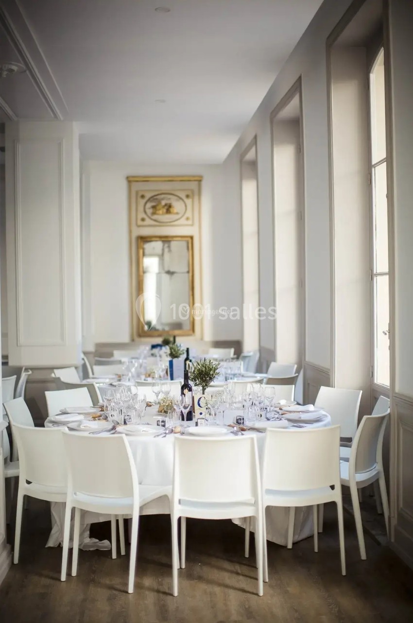 Salle lumineuse avec tables rondes dressées, chaises blanches et grand miroir doré en arrière-plan.