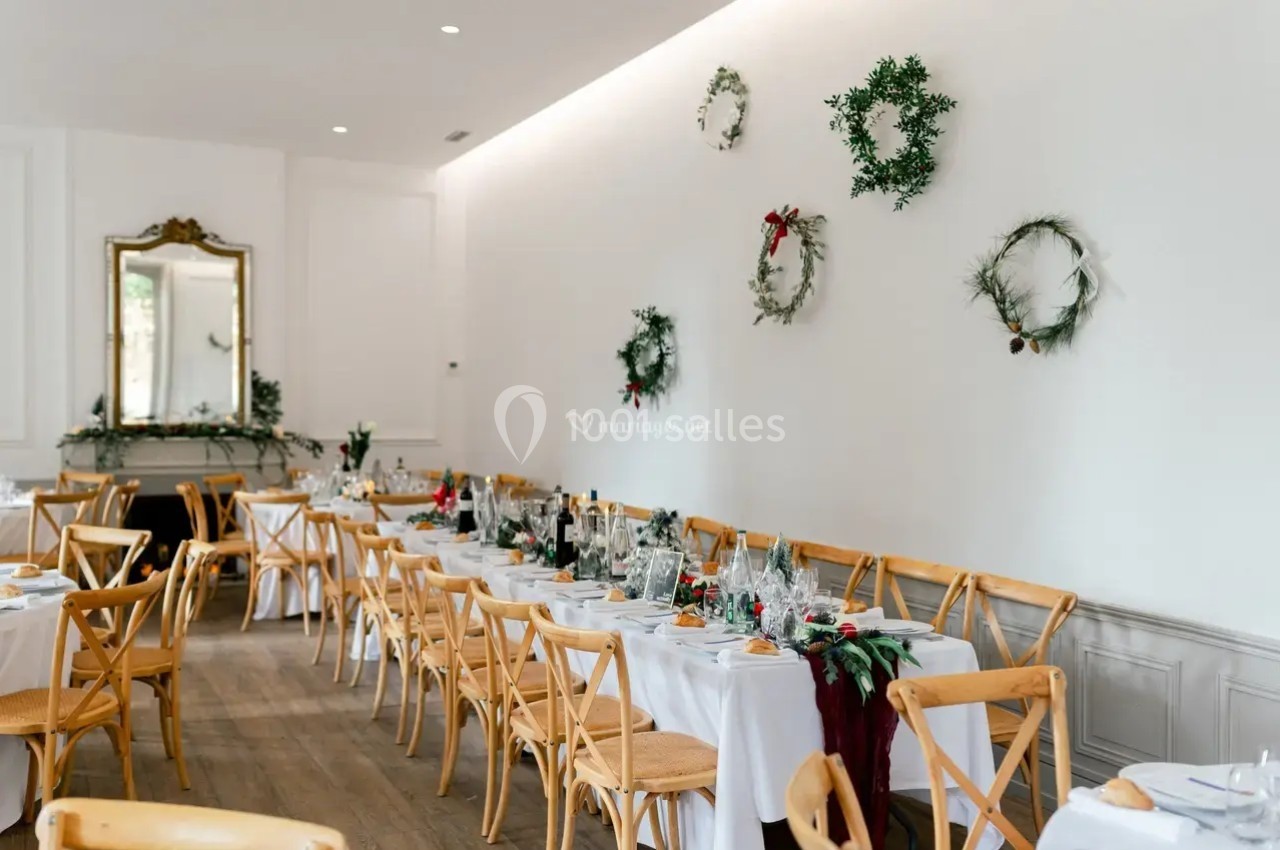 Salle de réception décorée pour un événement, avec tables dressées, chaises en bois et couronnes végétales au mur.