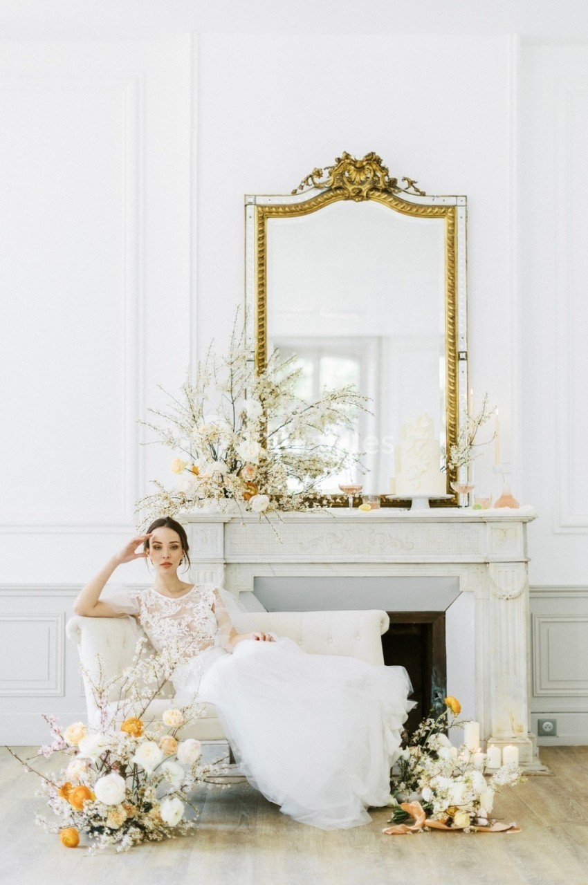 Femme en robe blanche assise sur un canapé devant une cheminée décorée de fleurs et d'un grand miroir doré.