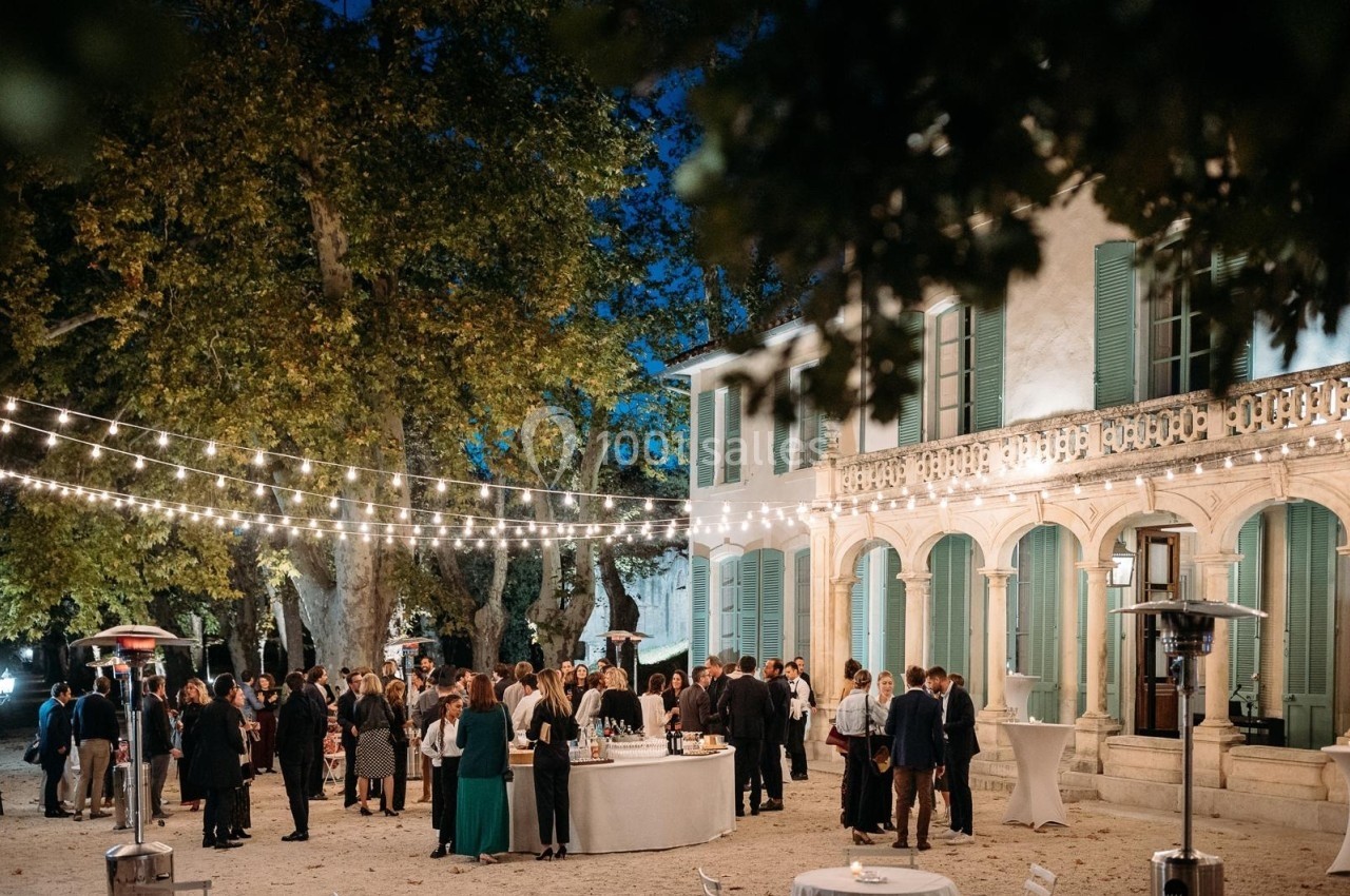 Groupe de personnes rassemblées en soirée dans une cour éclairée par des guirlandes lumineuses, devant un bâtiment ancien.