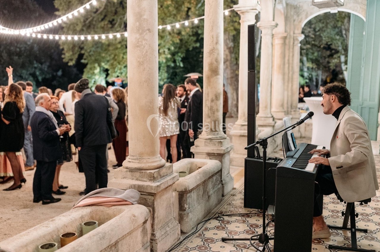 Un musicien joue du piano lors d'une réception en plein air, entouré d'invités discutant sous des guirlandes lumineuses.