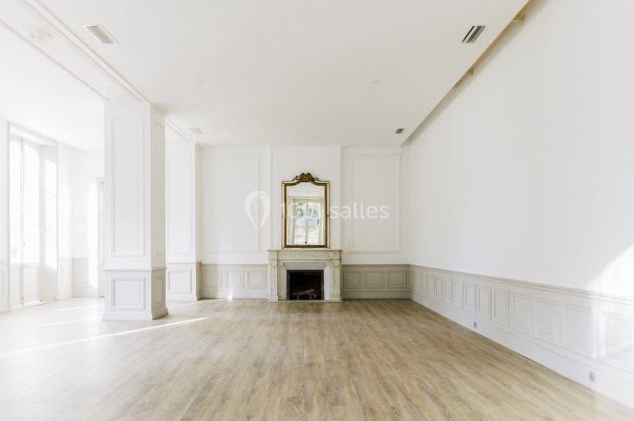 Salle lumineuse avec parquet clair, murs blancs, cheminée ornée d'un miroir doré et grandes fenêtres.