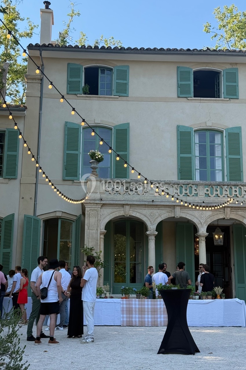 Groupe de personnes discutant devant une grande maison avec volets verts, décorée de guirlandes lumineuses.