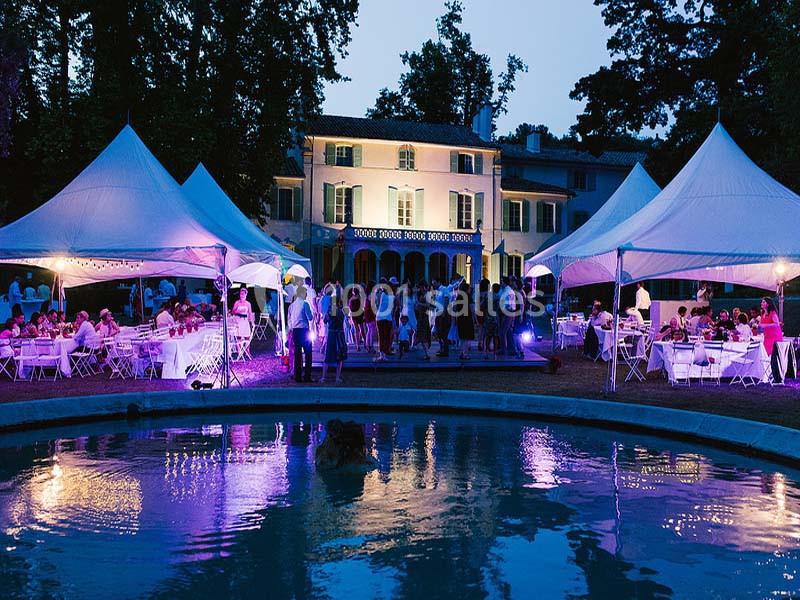 Soirée en plein air avec des invités sous des tentes blanches, devant une grande maison illuminée près d'un bassin.
