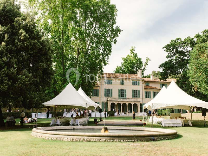 Grande maison entourée d’arbres avec des tentes blanches installées sur une pelouse et une fontaine au premier plan.