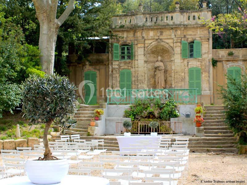Jardin avec des chaises blanches disposées devant une petite bâtisse ancienne ornée de volets verts et d'une statue.