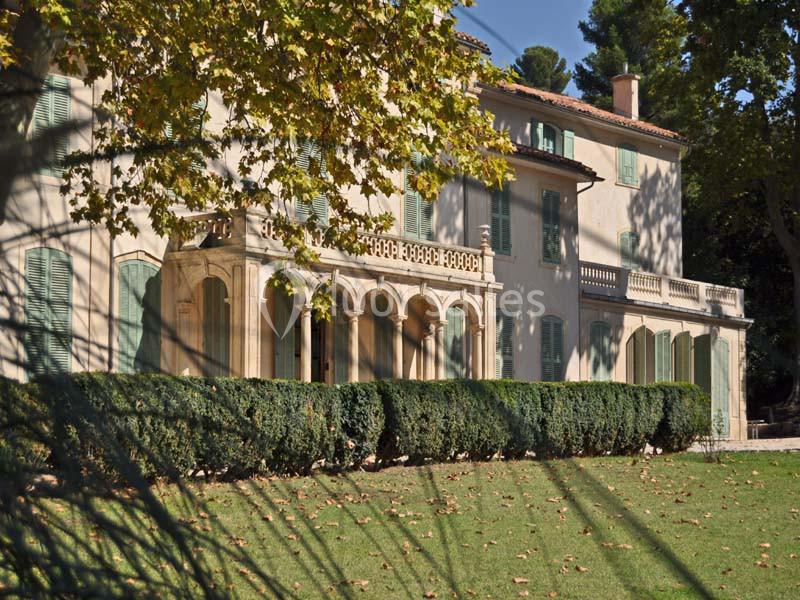 Façade d'une grande maison provençale entourée d'arbres, avec pelouse et ombres projetées au premier plan.