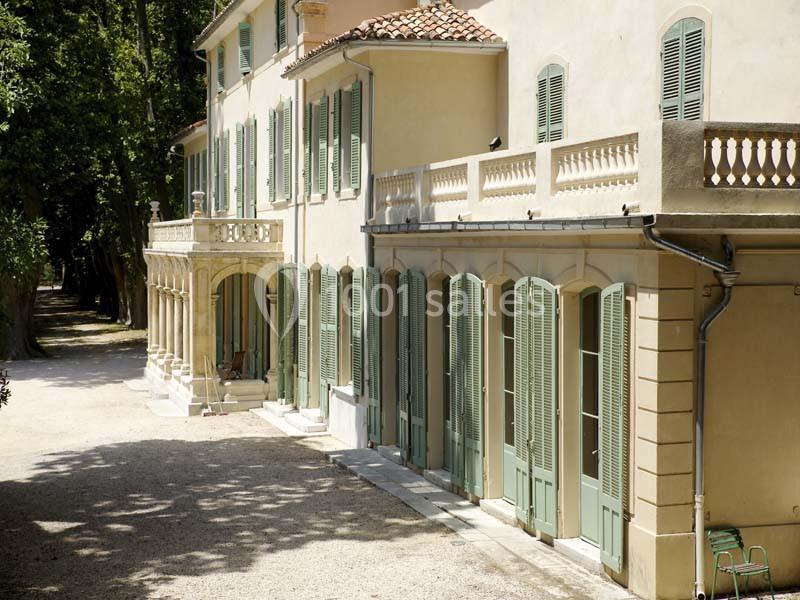 Façade d'une grande maison beige avec volets verts, balcons et allée ombragée par des arbres.