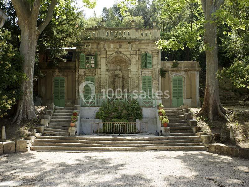 Façade d'un petit bâtiment en pierre entouré d'arbres, avec des volets verts et un escalier symétrique.