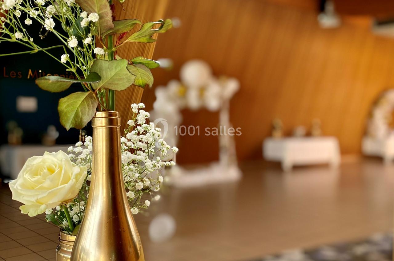 Centre de table avec bouteille dorée, fleurs blanches et jaunes, dans une salle en bois décorée pour un événement.