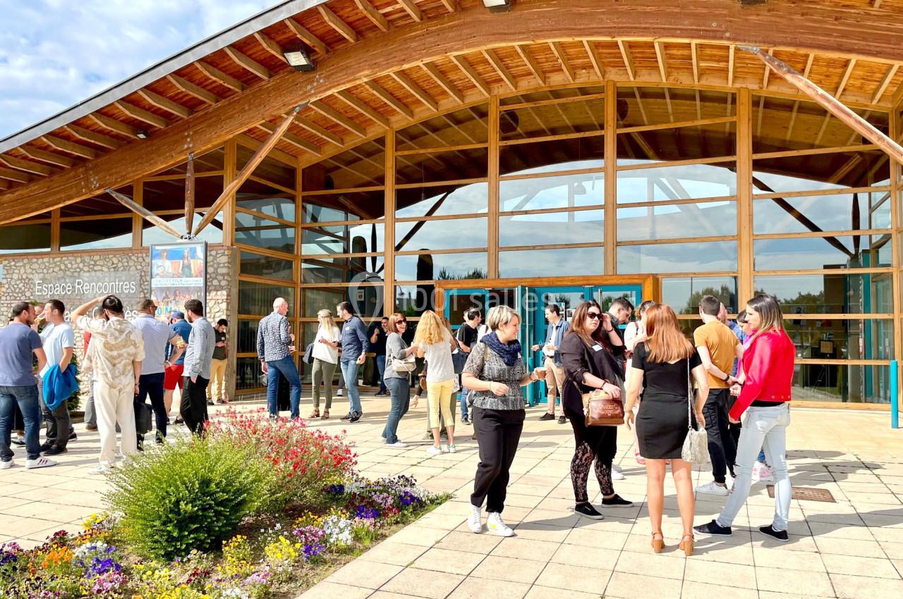 Groupe de personnes discutant devant un bâtiment en bois avec une façade vitrée, entouré de fleurs et d'une terrasse pavée.