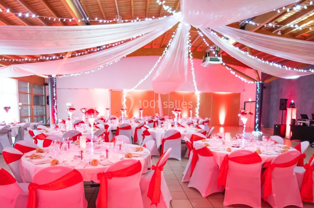 Salle de réception décorée avec des nappes blanches, des rubans rouges et des guirlandes lumineuses suspendues.