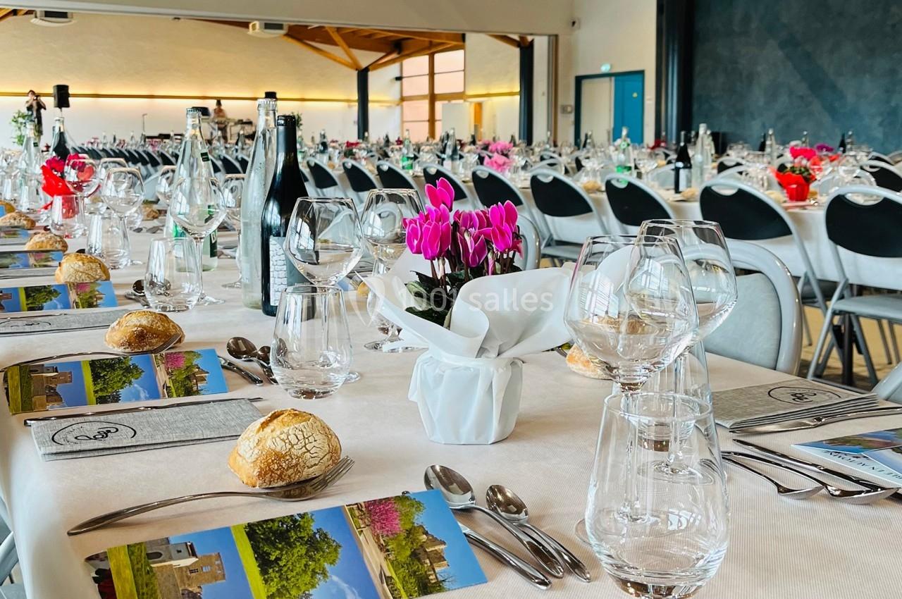 Tables dressées avec nappes blanches, couverts, verres, menus et décorations florales dans une grande salle lumineuse.