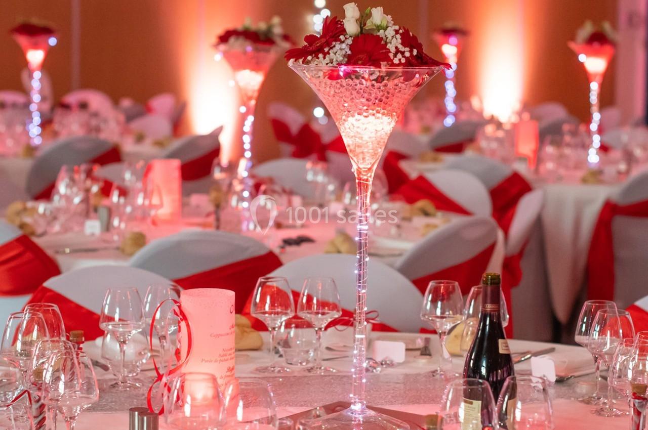 Salle de réception décorée avec des tables rondes, nappes blanches, nœuds rouges et centre de table lumineux.
